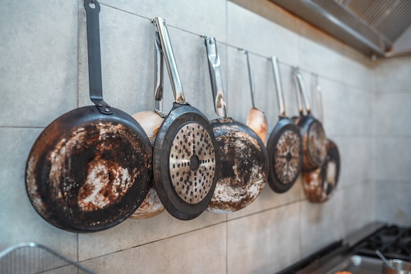 Indoor shot of a kitchen. Cooking equipment with carbon buildup on it hanging on the wall. Multiple frying pans with different dimensions.