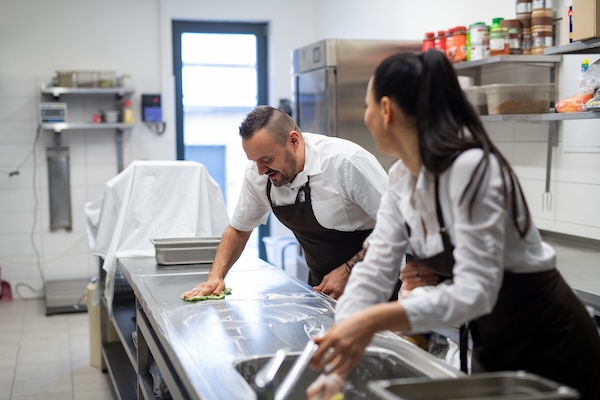 Two kitchen staff members use eco-friendly commercial cleaning products to sanitize a prep table