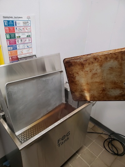 Someone loads a dirty sheet pan into an open FOG Tank, with a clean sheet pan ready to be removed from the tank