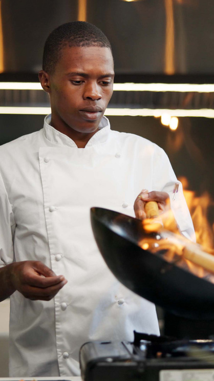 Black man, chef and fire with frying pan in kitchen at restaurant for fine dining, meal and creativity. Employee, professional and cooker with food for culinary, hospitality and restaurant service