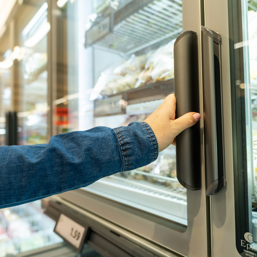 Hand opening a freezer door in a grocery store aisle, selecting refrigerated items