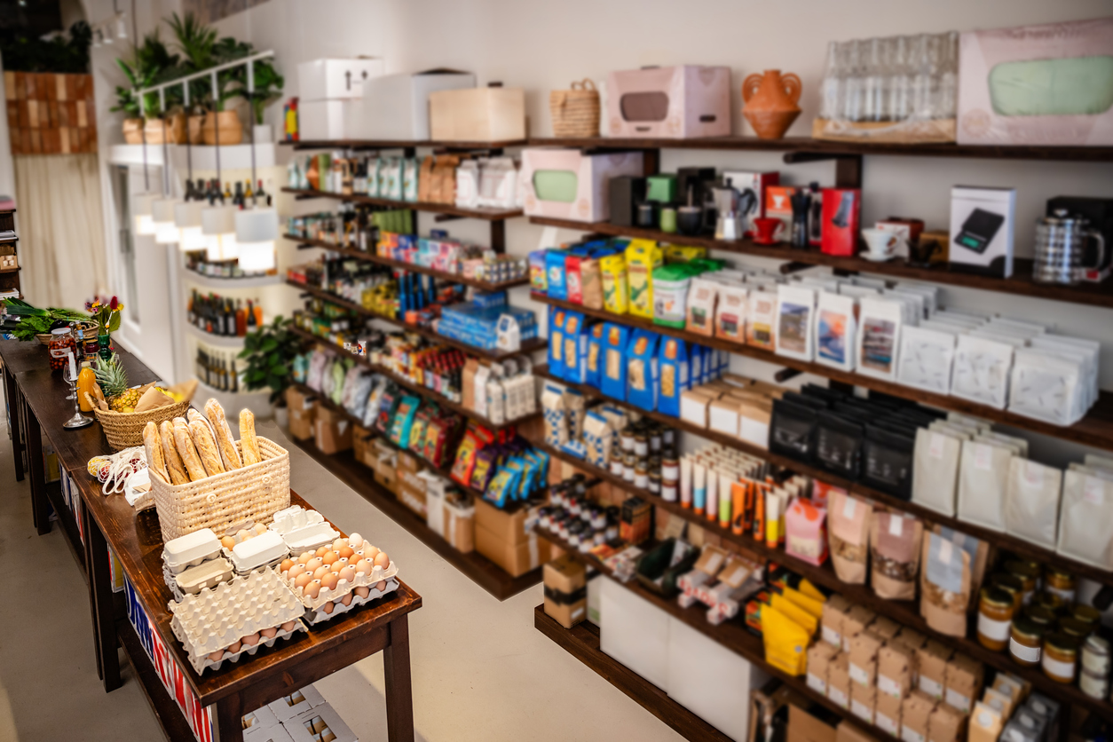 A vibrant grocery store aisle showcases a variety of products on wooden shelves. Items include packaged foods, beverages, and household goods, all arranged neatly. Natural light enhances the inviting atmosphere.